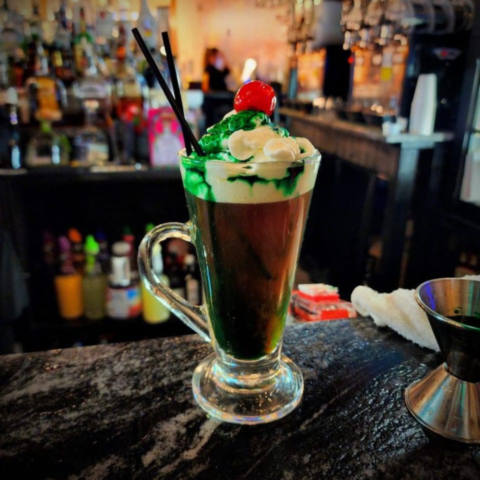 A green and white layered cocktail with a cherry and straws sits on a bar.