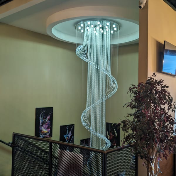 A large, modern spiral chandelier hangs from the ceiling above a railing, with cocktail art on the wall.