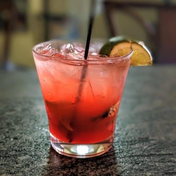 A vibrant red cocktail with ice, a black straw, and lime wedges on a dark speckled surface.