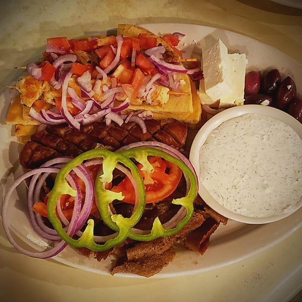 A large platter of Greek food with chicken skewers, gyro meat, feta, olives, and tzatziki sauce.