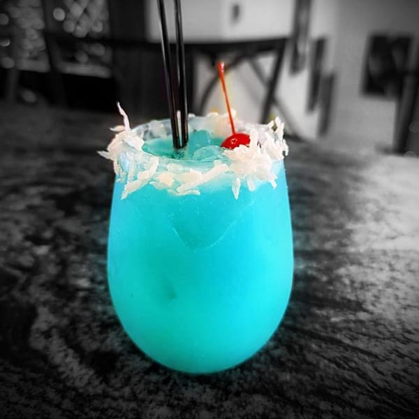 Vibrant blue cocktail with a coconut-flaked rim, two black straws, and a red cherry garnish.